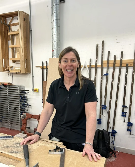 Smiling woman in a black polo shirt standing at a woodworking bench with tools and timber in a Glasgow Kelvin College workshop Smiling woman in a black polo shirt standing at a woodworking bench with tools and timber in a Glasgow Kelvin College workshop